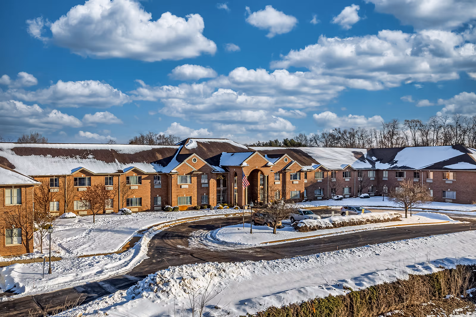 Exterior view of a large brick senior living facility building with a snow-covered roof and surrounding grounds. The building has multiple windows and a central entrance with an American flag in front. The sky is blue with scattered clouds.