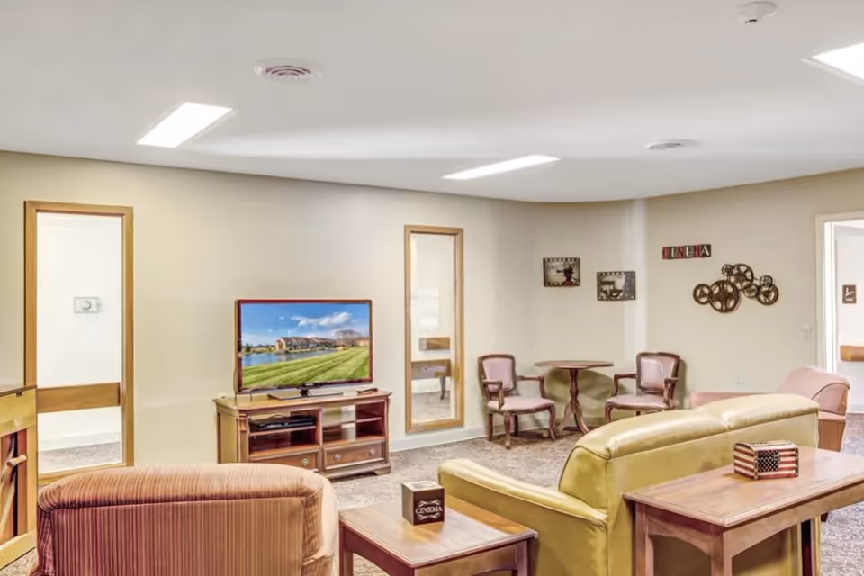 A cozy living room area in Reflections Retirement Community featuring a TV on a wooden stand, a green couch, a striped armchair, two wooden side tables with decorative boxes, and a small round table with two chairs. The walls are decorated with cinema-themed artwork and film reel decorations.