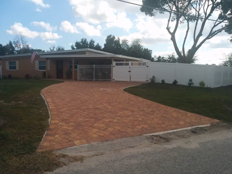 Front exterior of a single-story house with a brick paver driveway, white fence, and an American flag.