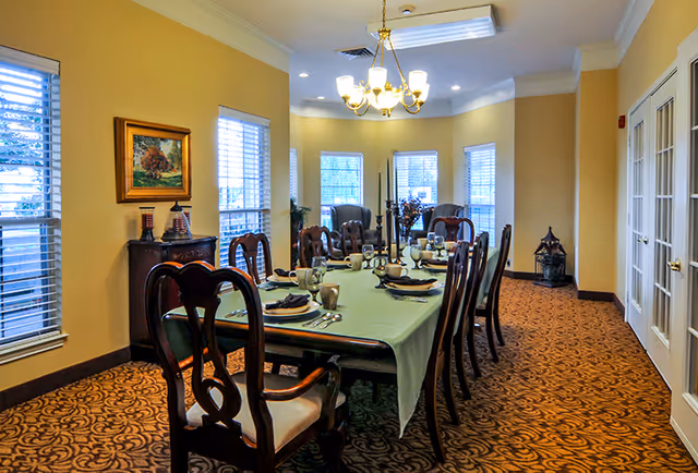 Formal dining room with a long table set for a meal, wooden chairs, chandelier, and windows along the walls.
