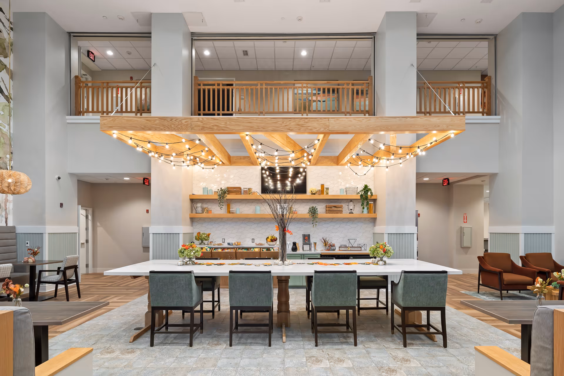 A spacious and modern common area in a senior living facility featuring a large white rectangular table with eight green upholstered chairs. Above the table is a wooden beam structure adorned with string lights. The back wall has two wooden shelves decorated with plants, jars, and other decorative items, and a flat-screen TV mounted in the center. The room has high ceilings with a balcony railing visible on the upper level. Additional seating areas with chairs and small tables are visible on either side of the main table.