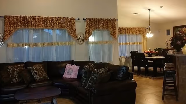 Interior view of a senior living facility showing a living room area with a dark leather sectional sofa adorned with multiple patterned cushions and a pink pillow. The windows have sheer white curtains with decorative brown and gold valances. Adjacent to the living room is a dining area with a dark wooden dining table and chairs, a hanging light fixture, and floral decorations on the table and side surfaces.