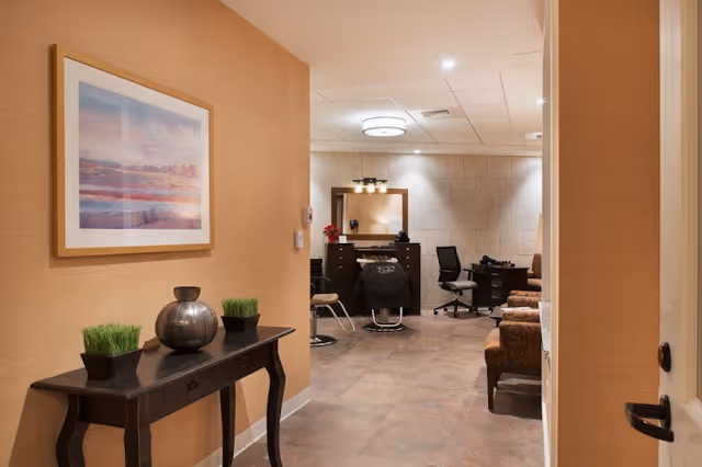 Interior view of a salon area in a senior living facility with a styling chair, mirror, and workstation. The foreground shows a hallway with a black console table decorated with two small potted plants and a silver vase, and a framed landscape painting on the wall.
