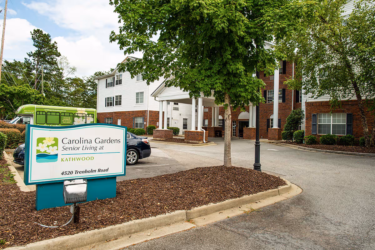 Entrance to Carolina Gardens at Kathwood senior living facility showing a white and brick building with a covered driveway, a tree in the foreground, parked cars, and a sign displaying the facility name and address at 4520 Trenholm Road.