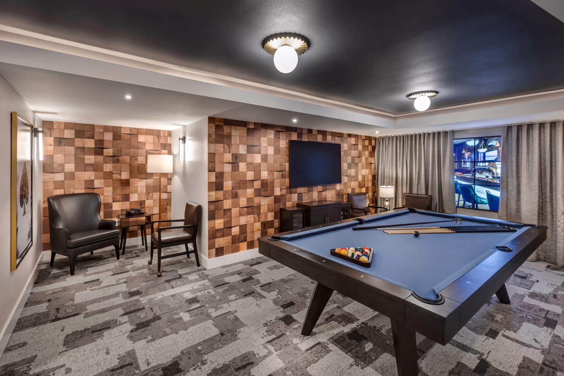 A modern game room with a pool table in the center, a flat-screen TV mounted on a wooden textured wall, two chairs and a small table with a lamp in the corner, and large curtains covering a window.