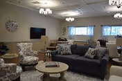 A cozy living room area in a senior living facility with a dark gray sofa, patterned armchairs, round wooden coffee tables, a wall-mounted flat screen TV, large windows with curtains, and ceiling lights.