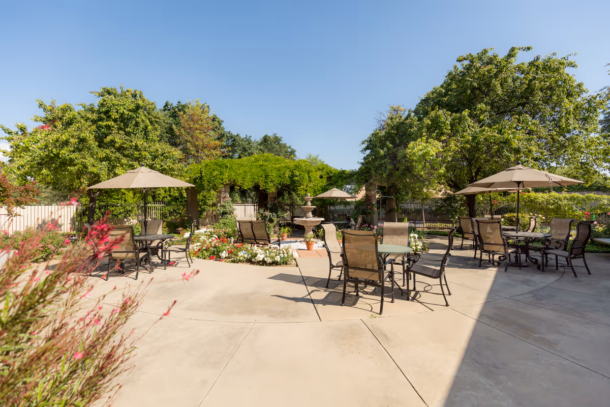 Outdoor patio area with multiple tables and chairs under large umbrellas, surrounded by green trees, flowering plants, and a central water fountain under a clear blue sky.