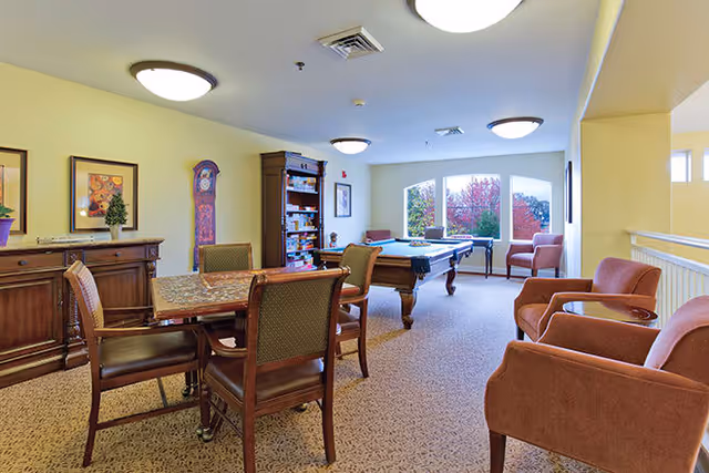 A well-lit common area in a senior living facility featuring a pool table, a table with four chairs, several armchairs, a wooden cabinet, a bookshelf, and large windows showing colorful autumn trees outside.