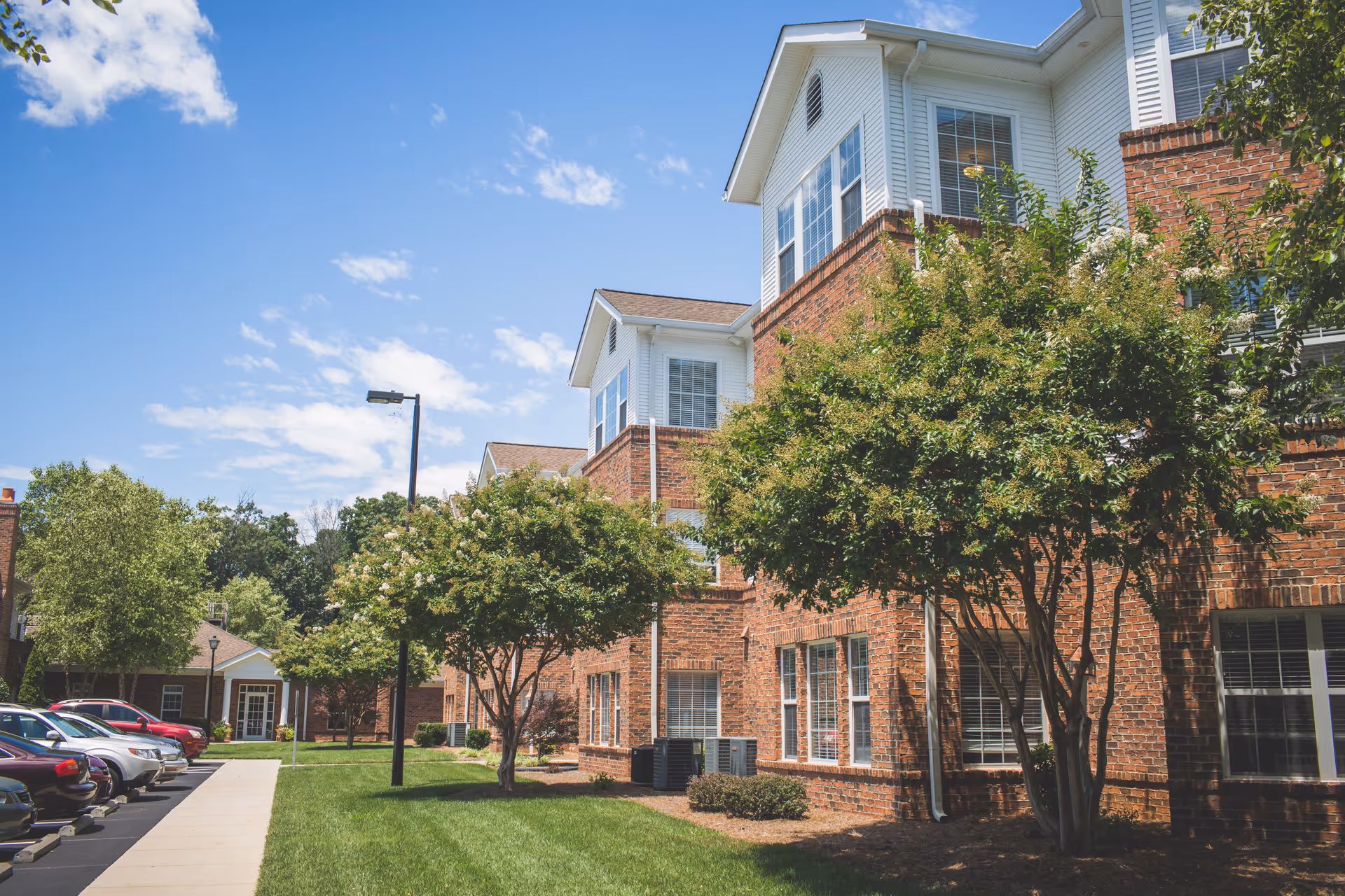 Exterior view of a senior living facility named Homestead Hills showing a brick building with white trim, multiple windows, and a well-maintained lawn with trees and a sidewalk. Several parked cars are visible along the left side under a blue sky with scattered clouds.