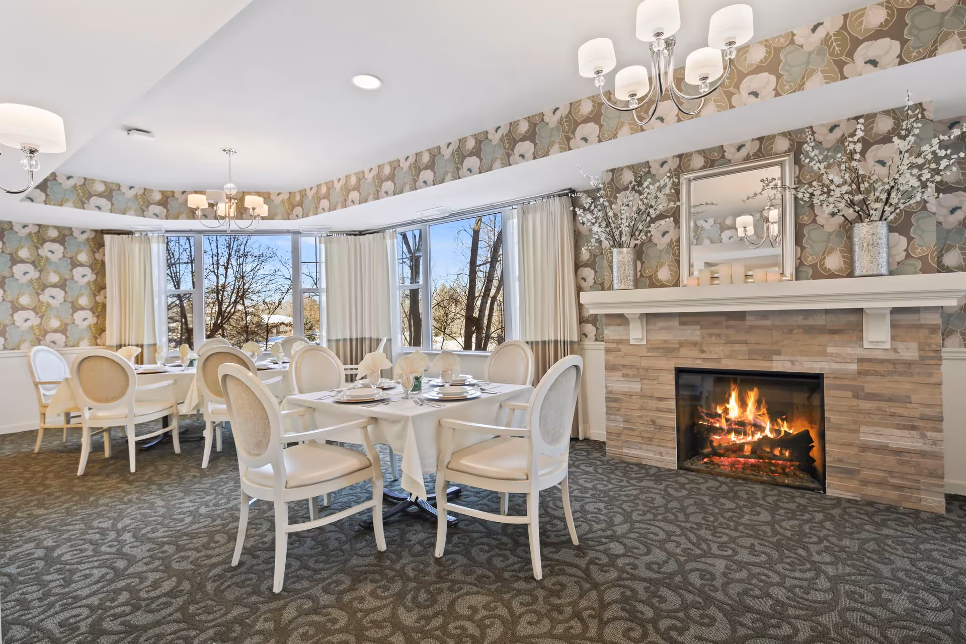 A cozy dining room with round tables covered in white tablecloths and set with plates and napkins. The room features white cushioned chairs, a lit fireplace with a stone surround, floral wallpaper, and large windows with cream-colored curtains letting in natural light. Decorative vases with white flowers and a mirror are placed on the mantel above the fireplace.