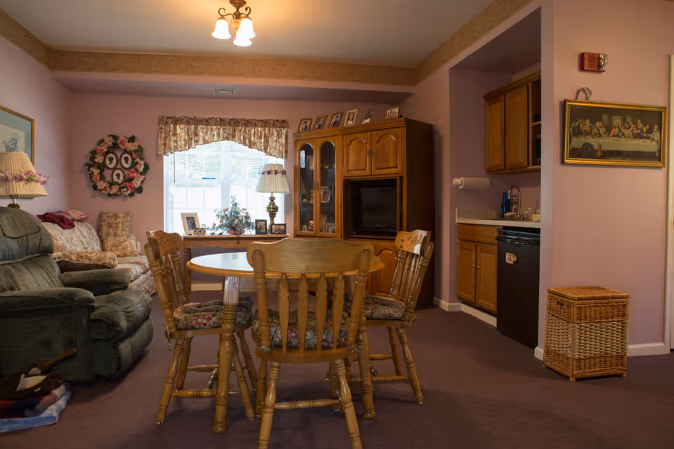 A cozy senior living room with a round wooden dining table and chairs, upholstered recliner and sofa, TV cabinet and a small kitchenette.