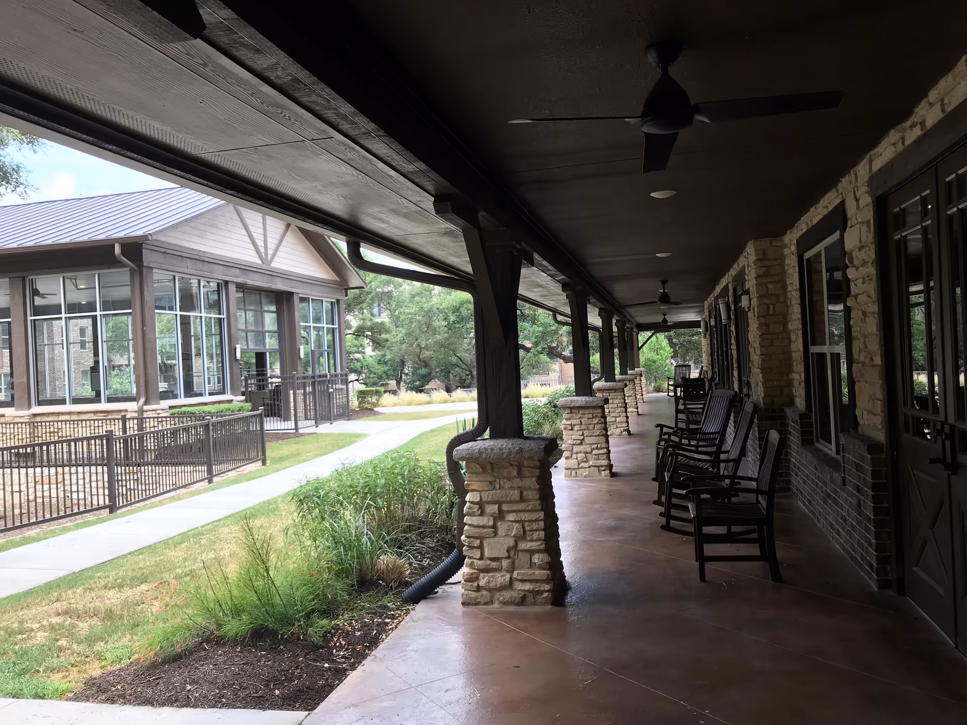 Covered outdoor porch area with stone pillars and several rocking chairs lined up along the wall of a building. The porch overlooks a landscaped garden area with grass and shrubs, and another building with large windows is visible across a paved walkway.