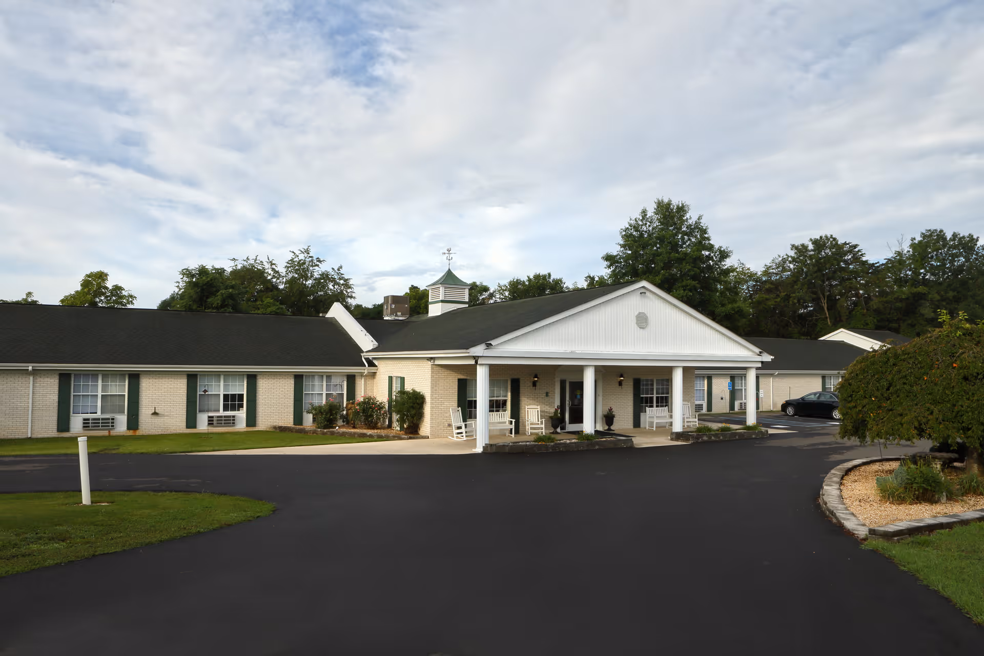 Single-story light-brick senior living building with a covered entrance supported by white columns, rocking chairs on the porch, a curved driveway and landscaping under a cloudy sky.
