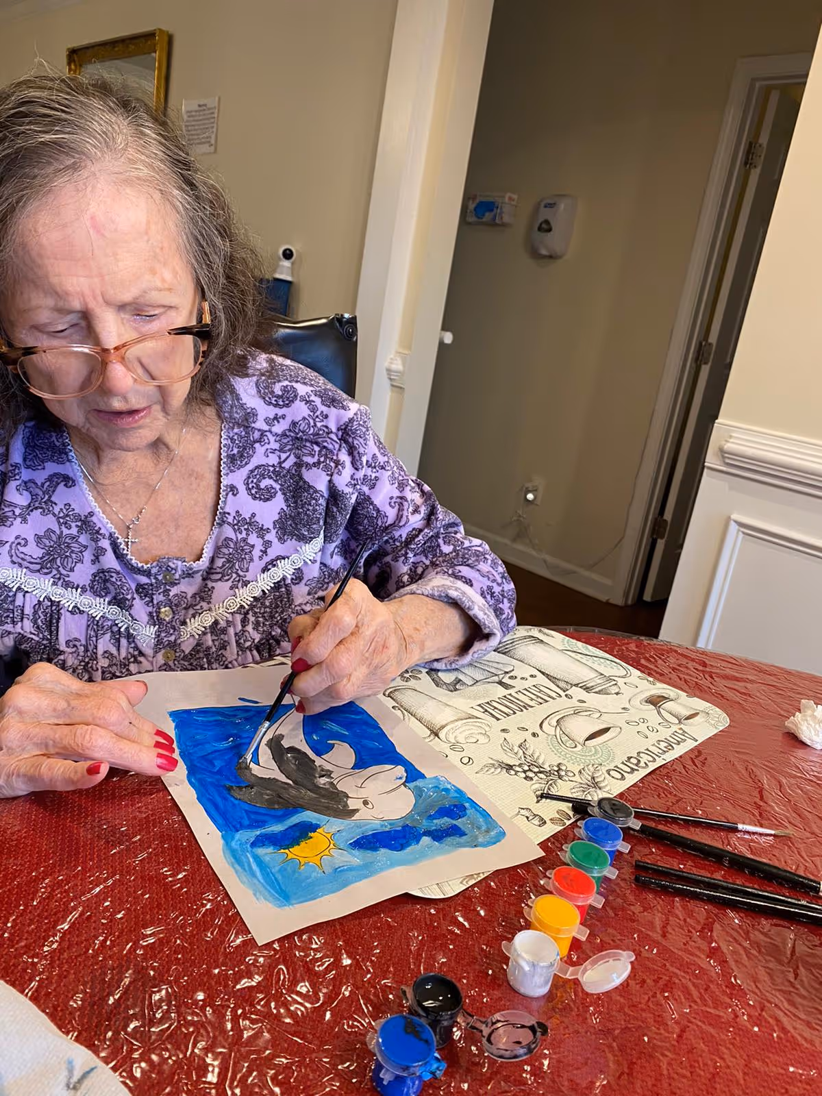 An elderly woman wearing glasses and a purple patterned top is painting a colorful picture of a dolphin jumping out of the water with a sun in the background. She is seated at a table covered with a red plastic tablecloth, with several small paint containers and brushes laid out in front of her.