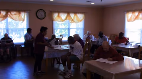 Residents and staff gathered in a dining/activity room, seated at tables and filling out papers.