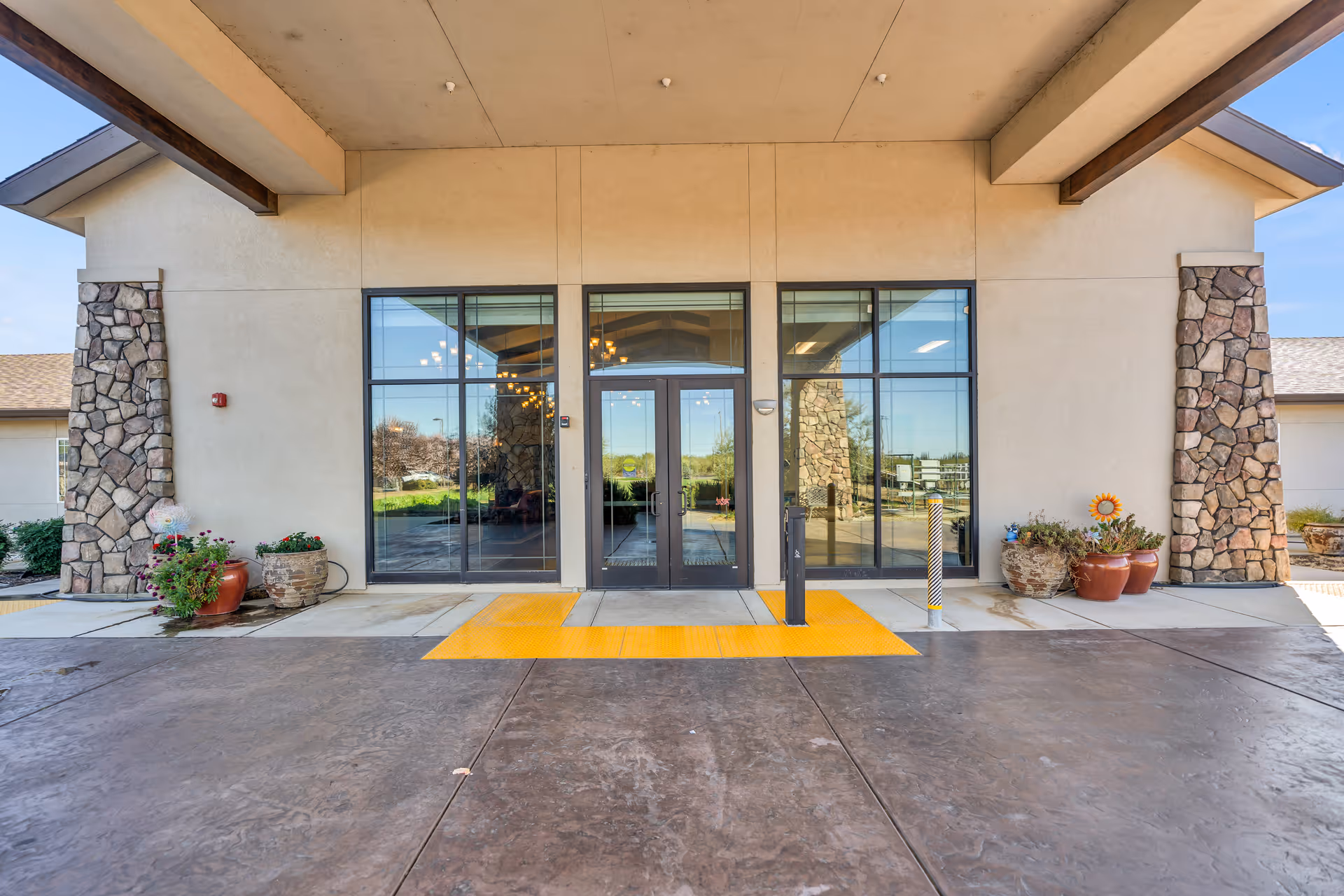 Covered entrance to Emerald Oaks Inc with glass double doors flanked by stone columns, potted plants, and a yellow tactile walkway.