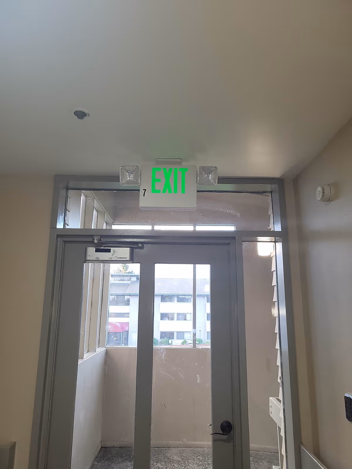 Interior view of an exit door with a green illuminated EXIT sign above it. The door has glass panels and leads to an outdoor balcony area with a view of a building across the way. The walls are beige, and there is a smoke detector on the ceiling and a keypad on the right wall.