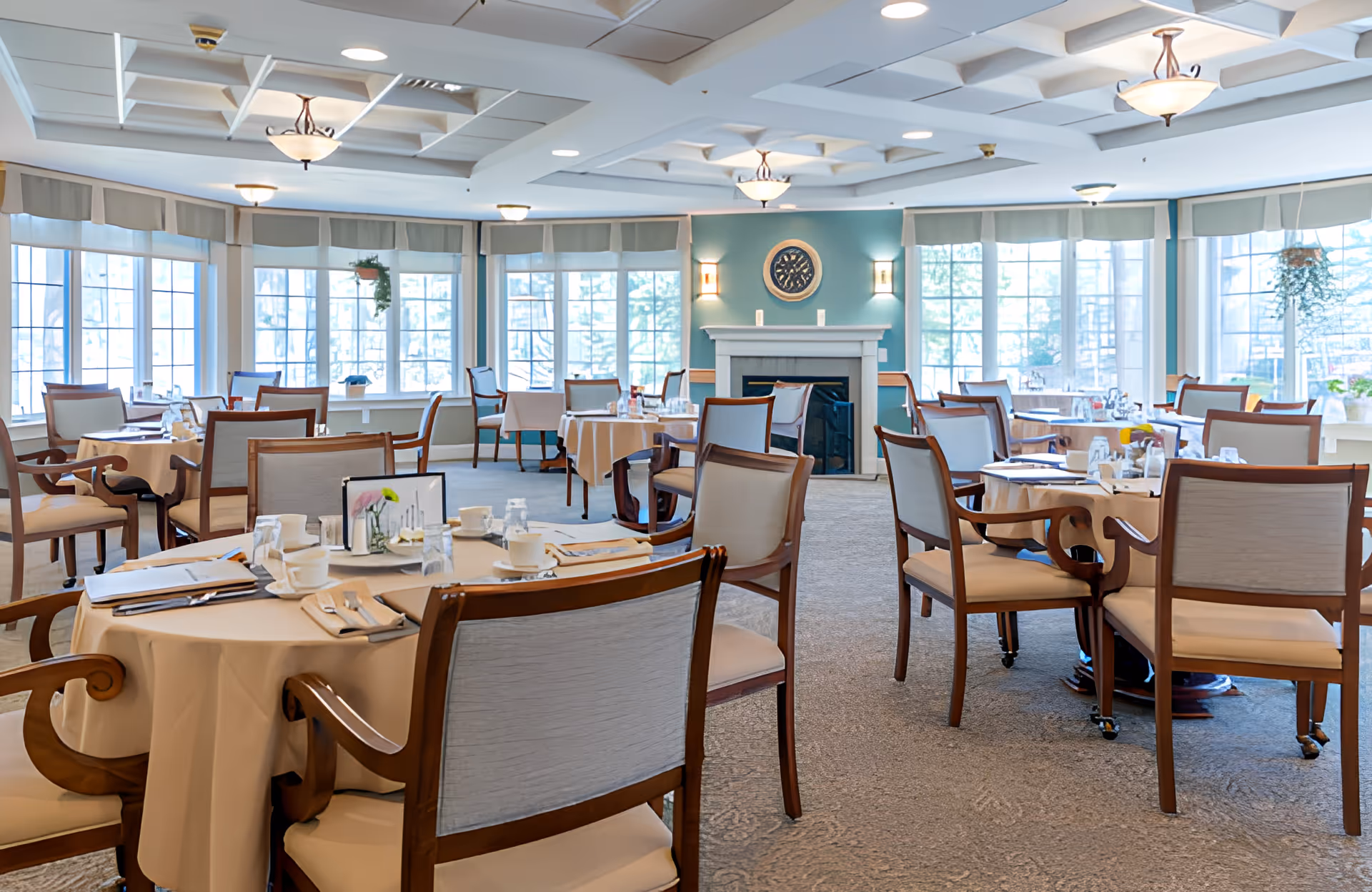 Bright, spacious dining room with round tables set for meals, wooden chairs, large windows and a fireplace.