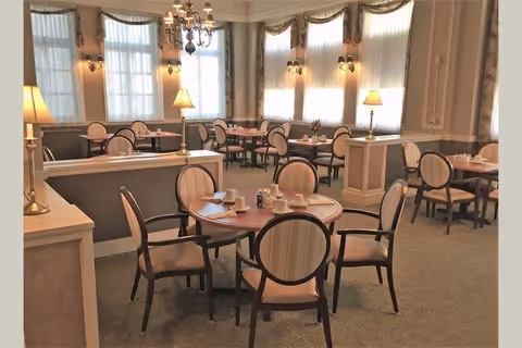 Elegant dining room with round tables set for meals, upholstered chairs, chandeliers, lamps, and large curtained windows.