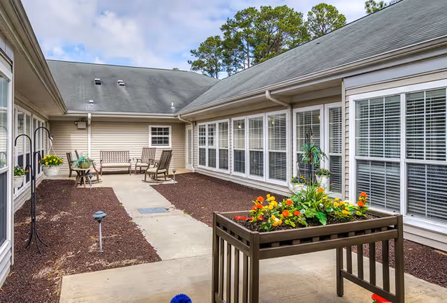 Outdoor courtyard area with a concrete walkway, brown mulch beds, and a raised planter box filled with colorful flowers. There are several wooden chairs and a bench along the building walls with large windows and white blinds. The building has beige siding and a gray roof under a partly cloudy sky.