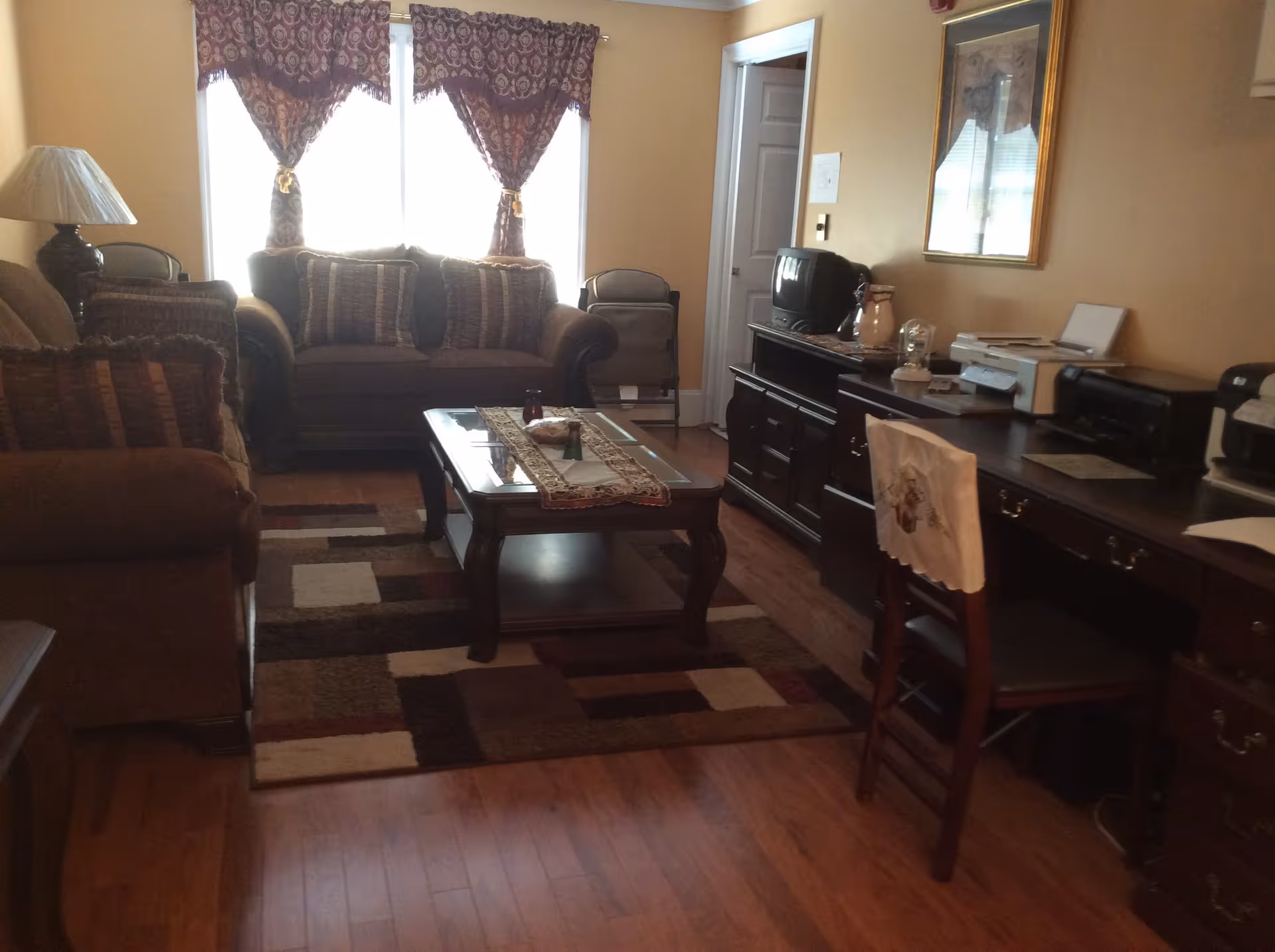 A cozy living room with two brown sofas, a wooden coffee table with a decorative runner, a patterned area rug, and a wooden desk with a chair. The room has wooden flooring, a table lamp, and windows with patterned curtains letting in natural light.