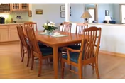 A dining area with a wooden table and six matching wooden chairs with dark cushions. The table has a floral centerpiece and a lace doily. In the background, there is a kitchen area with wooden cabinets, a countertop, and a window opening to the dining room. The floor is hardwood, and the walls are light-colored.