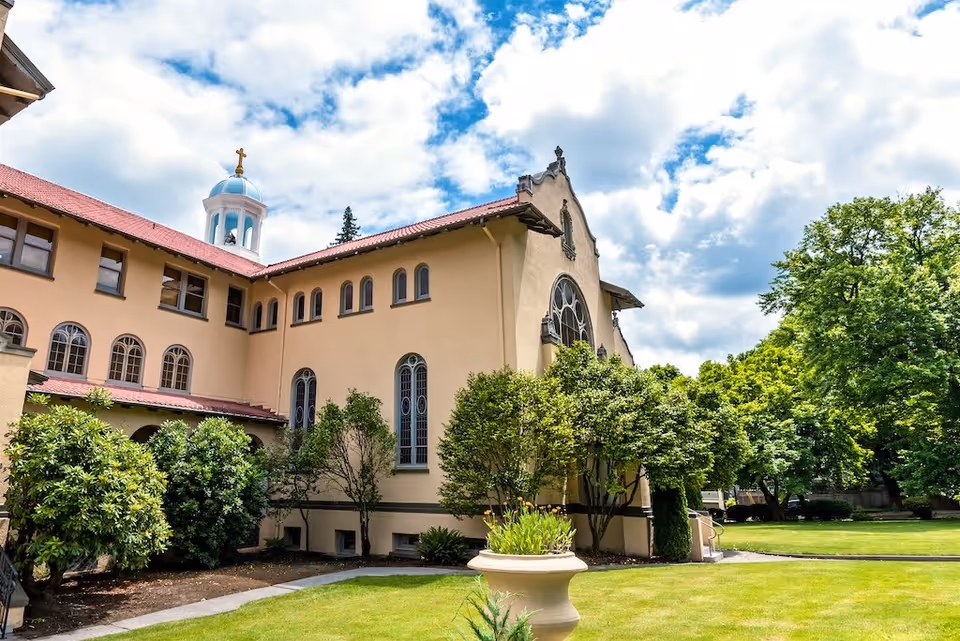 Exterior view of a large beige building with arched windows and a red roof, surrounded by green trees and a well-maintained lawn under a partly cloudy blue sky.