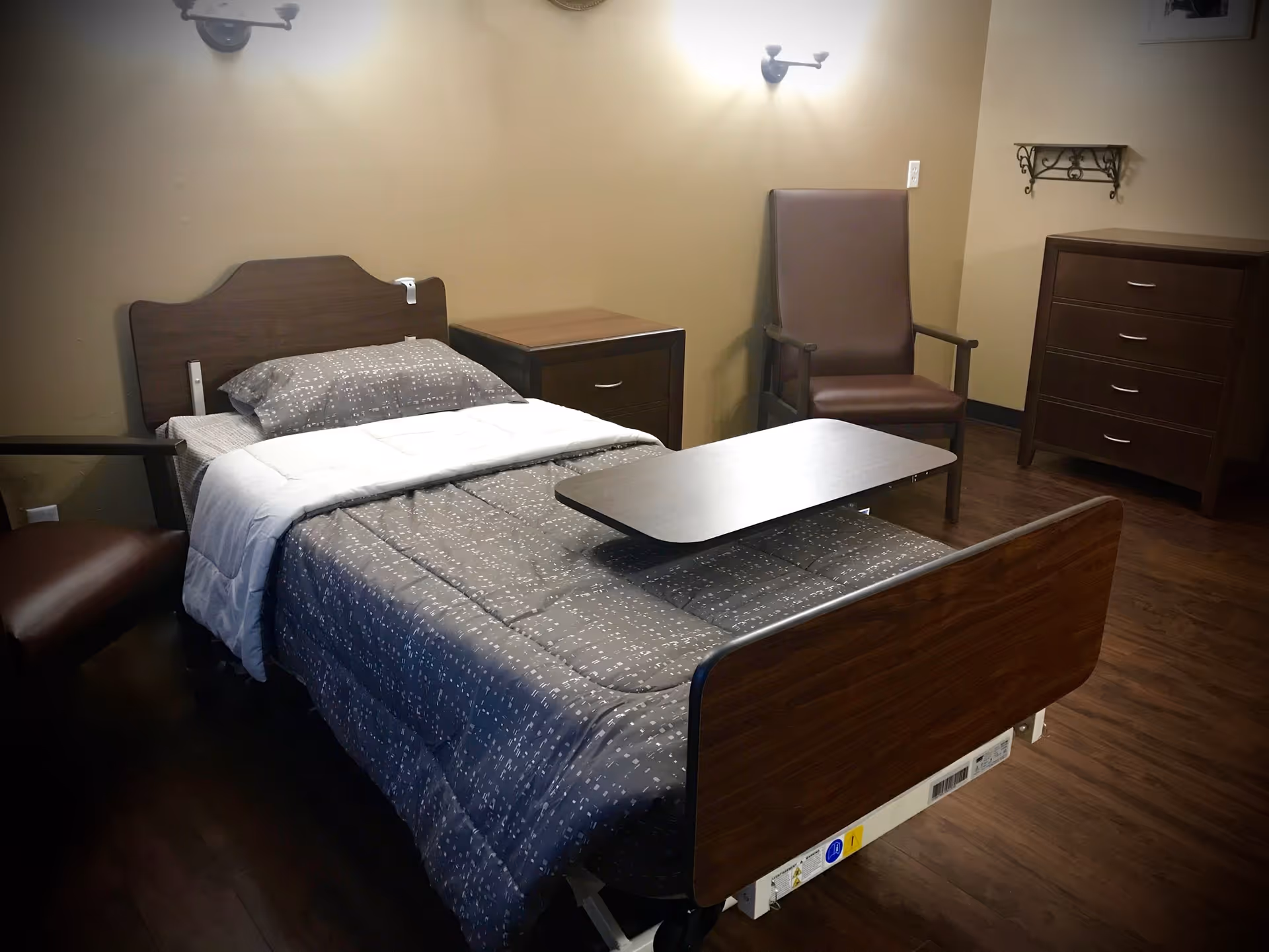 A senior living facility bedroom with a single hospital-style bed covered in a gray patterned comforter and white blanket. The room includes a wooden nightstand, a brown armchair, a wooden dresser, and a small wall-mounted shelf. The walls are painted beige and the floor is dark wood.