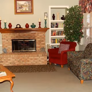 Cozy living room with a brick fireplace, a wooden mantel decorated with candles and small vases, a red armchair with a cushion, a patterned sofa, a built-in bookshelf filled with books and decorative items, a large green plant, and a window with floral curtains.