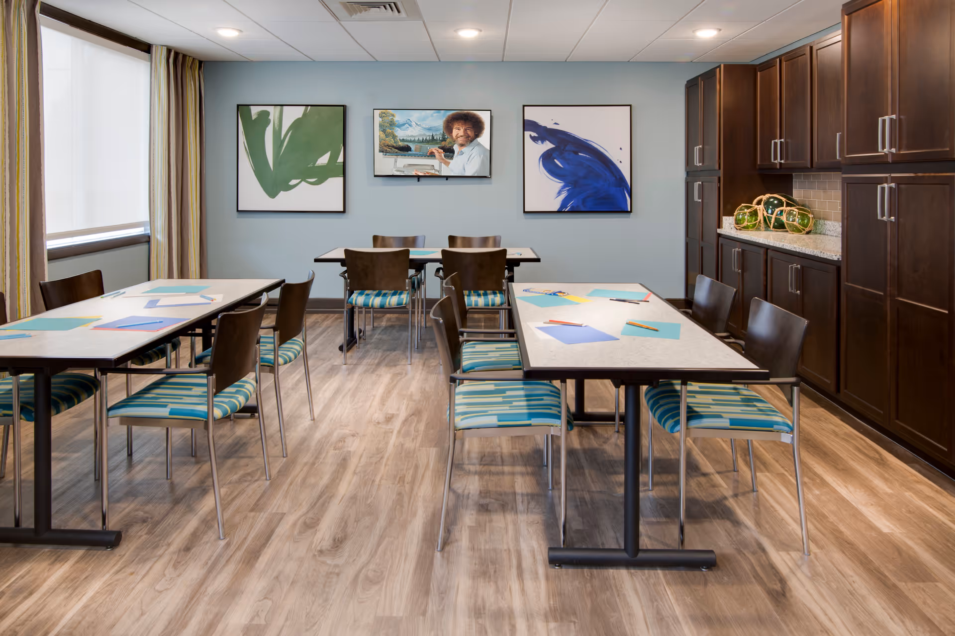 A well-lit activity or meeting room with two rectangular tables surrounded by chairs with blue and green striped cushions. The tables have colored paper and pencils on them. The room has light wood flooring, light blue walls, and dark wood cabinets along one wall. Three framed pictures hang on the wall, including one of Bob Ross painting a landscape. A large window with curtains is on the left side.