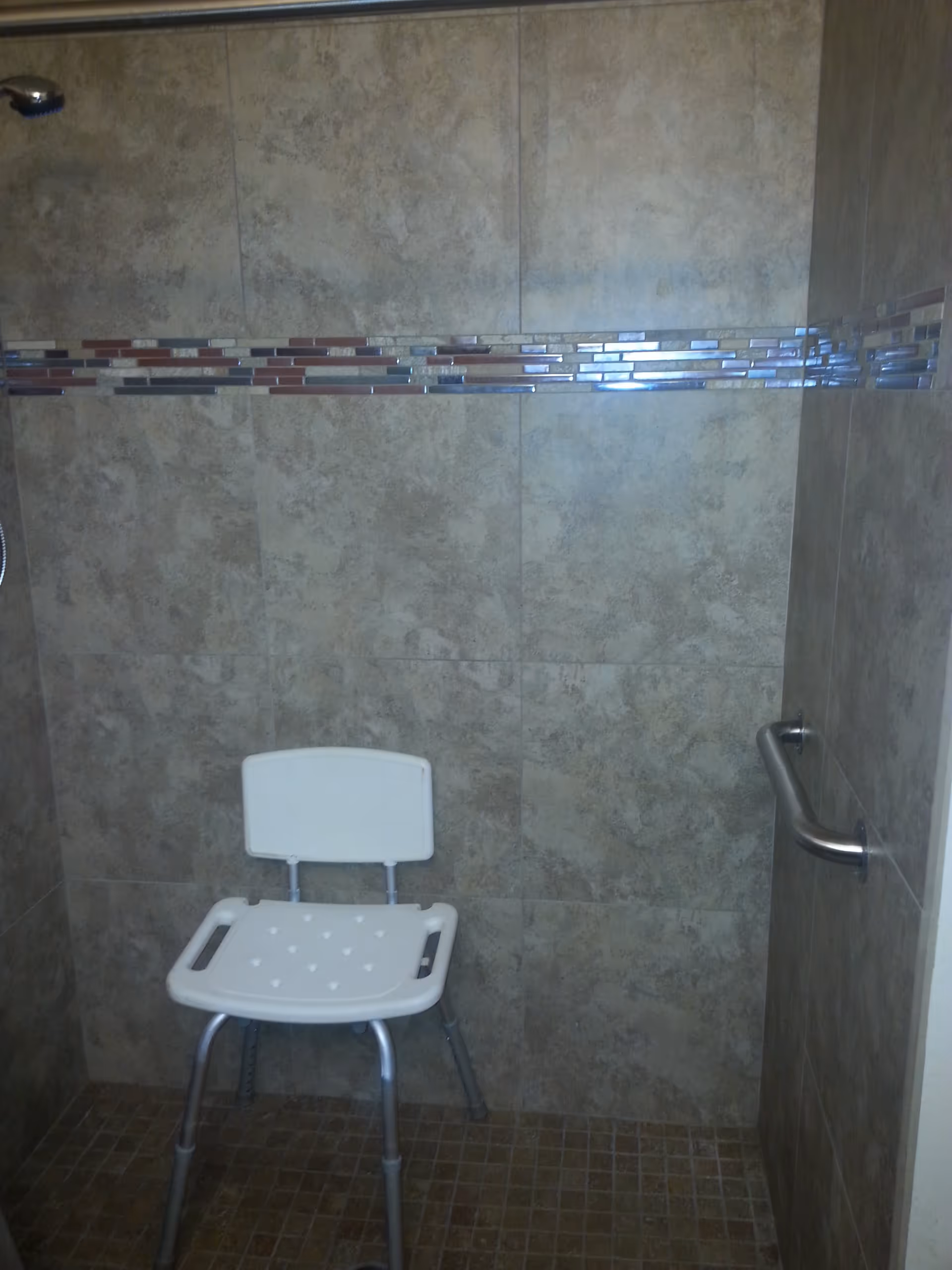 Shower area with beige tiled walls and floor, featuring a white plastic shower chair with metal legs and a metal grab bar mounted on the right wall.