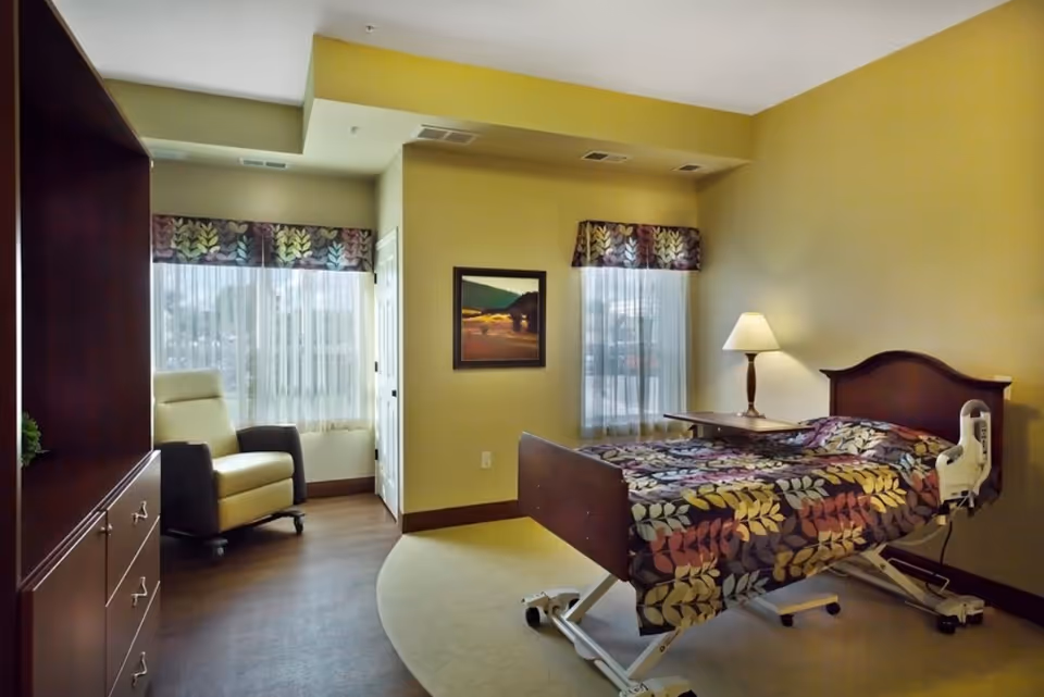 A senior living facility bedroom with a hospital-style adjustable bed covered in a colorful leaf-patterned bedspread, a wooden nightstand with a lamp, a comfortable beige and brown armchair near two windows with sheer curtains and patterned valances, and a wooden dresser with drawers.