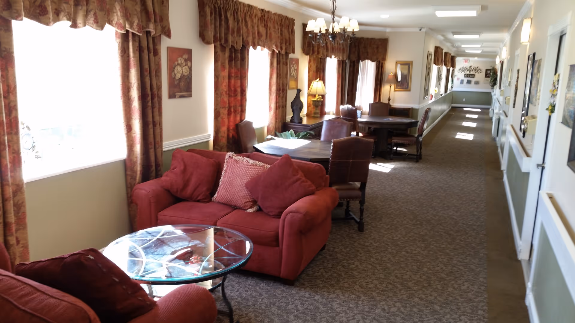 A cozy interior hallway area with red upholstered armchairs and a matching loveseat around a glass coffee table. There are several wooden tables with chairs along the wall, decorated with floral curtains, framed artwork, and lamps. The hallway extends into the distance with carpeted flooring and light-colored walls.