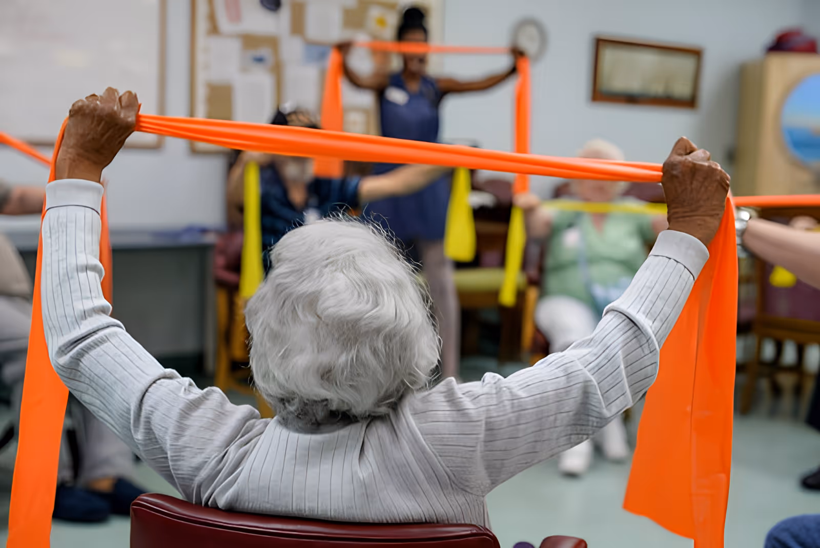 A group of elderly individuals seated in a room participating in a seated exercise class using colorful resistance bands, led by an instructor standing in the background.