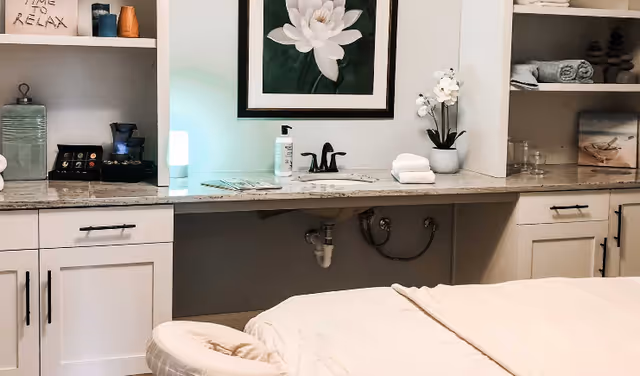 A serene spa or massage room with a massage table covered in a beige sheet in the foreground. Behind the table is a countertop with a sink, hand sanitizer, folded towels, a potted orchid, and shelves holding neatly folded towels and decorative items. A framed picture of a white flower hangs on the wall above the sink.
