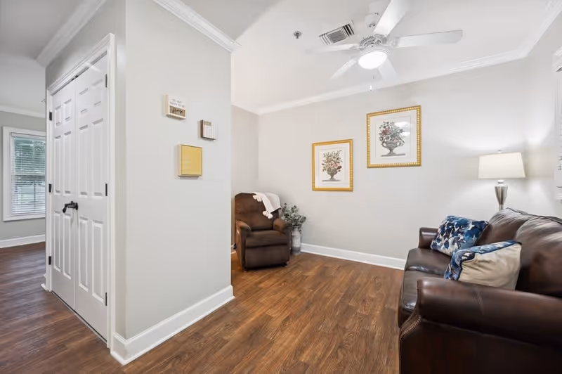 A cozy living room area with a brown leather sofa adorned with blue and white pillows, a brown recliner chair with a white throw draped over it, two framed floral prints on the wall, a floor lamp with a white shade, and wood flooring. There is a white closet with double doors on the left side and a window with blinds in the background.
