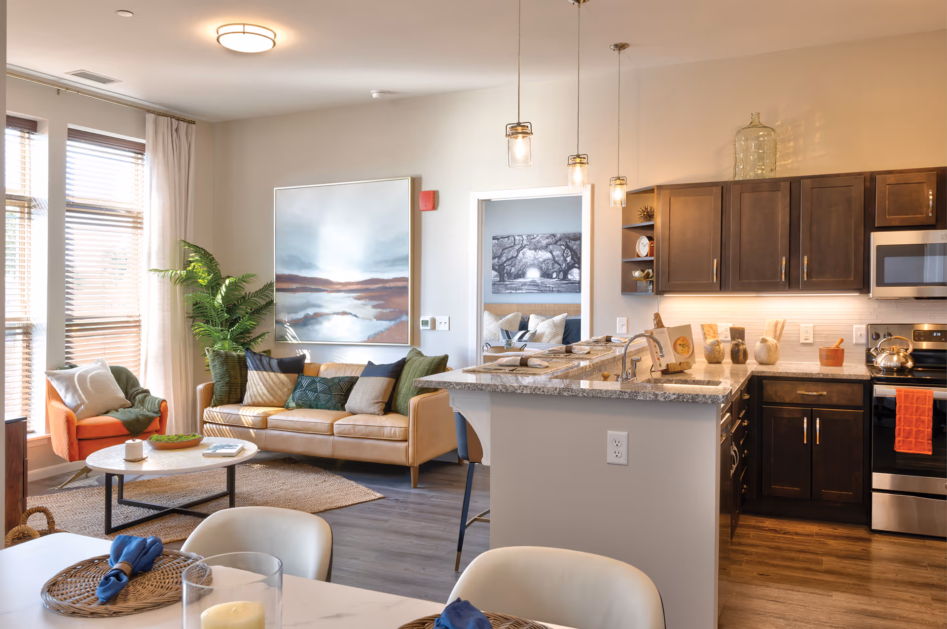 A modern open-concept living area featuring a tan leather sofa with multiple cushions, an orange armchair, a round coffee table, and a large window with blinds and curtains. Adjacent to the living area is a kitchen with dark wood cabinets, granite countertops, stainless steel appliances, and pendant lights hanging above the kitchen island. A dining table with white chairs and place settings is partially visible in the foreground. A doorway leads to a bedroom with a large black and white tree photograph above the bed.