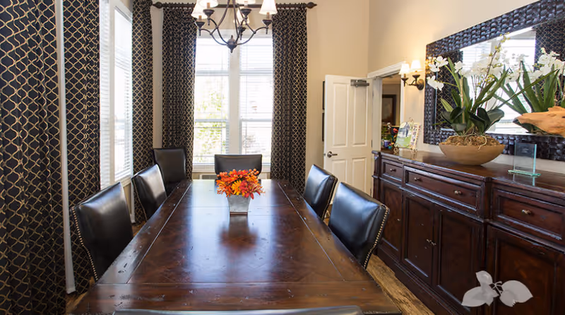 A well-lit dining room with a long wooden table surrounded by six black leather chairs. The table has a small vase with orange flowers in the center. Large windows with patterned dark curtains allow natural light to fill the room. On the right side, there is a wooden sideboard with a large decorative mirror above it and a potted plant with white flowers. A chandelier hangs from the ceiling above the table.