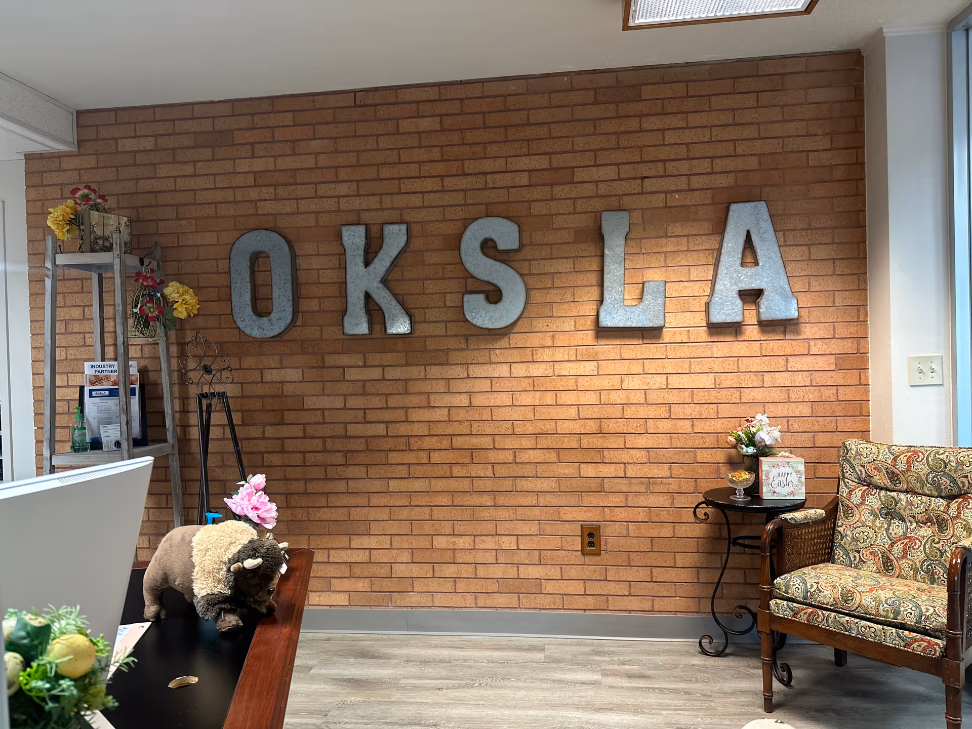 Interior view of a senior living facility reception or waiting area with a brick wall displaying large metal letters spelling 'OKSLA'. The room features a patterned armchair, a small round table with flowers and a decorative sign, a shelf with flowers and documents, and a plush buffalo toy on a counter.