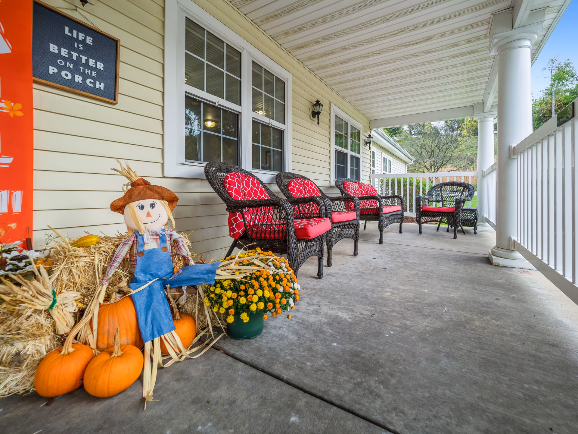 A covered porch with a row of black wicker chairs with red cushions along the wall of a light yellow building. On the left side, there is a fall-themed decoration including a scarecrow, pumpkins, hay bales, and a pot of yellow flowers. A sign on the wall reads 'Life is better on the porch.' The porch has white railings and columns, and trees are visible in the background.
