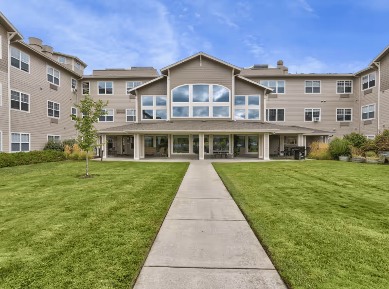 Front exterior of a multi-story senior living facility with a central entrance, large windows, and a well-kept green lawn.