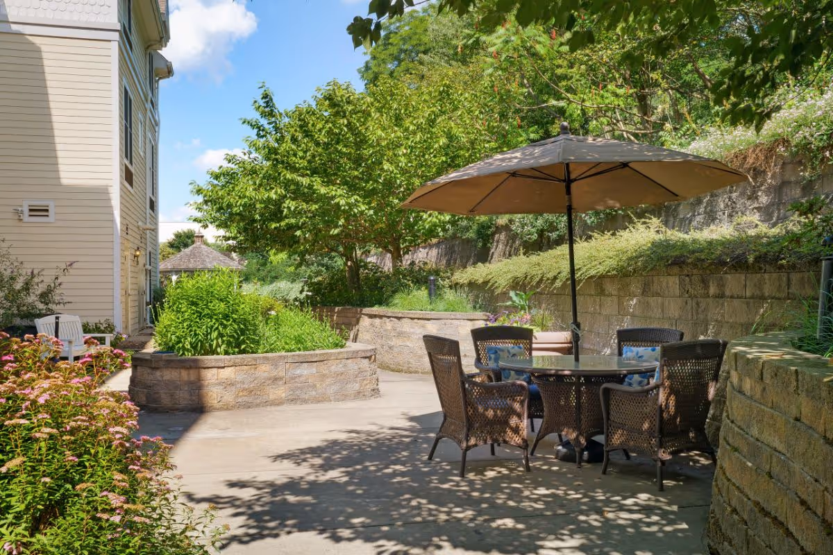 Outdoor patio area with a round glass table surrounded by four wicker chairs with cushions, shaded by a large beige umbrella. The patio is bordered by stone retaining walls and lush greenery including trees and flowering plants. A beige building is visible on the left side under a blue sky with some clouds.