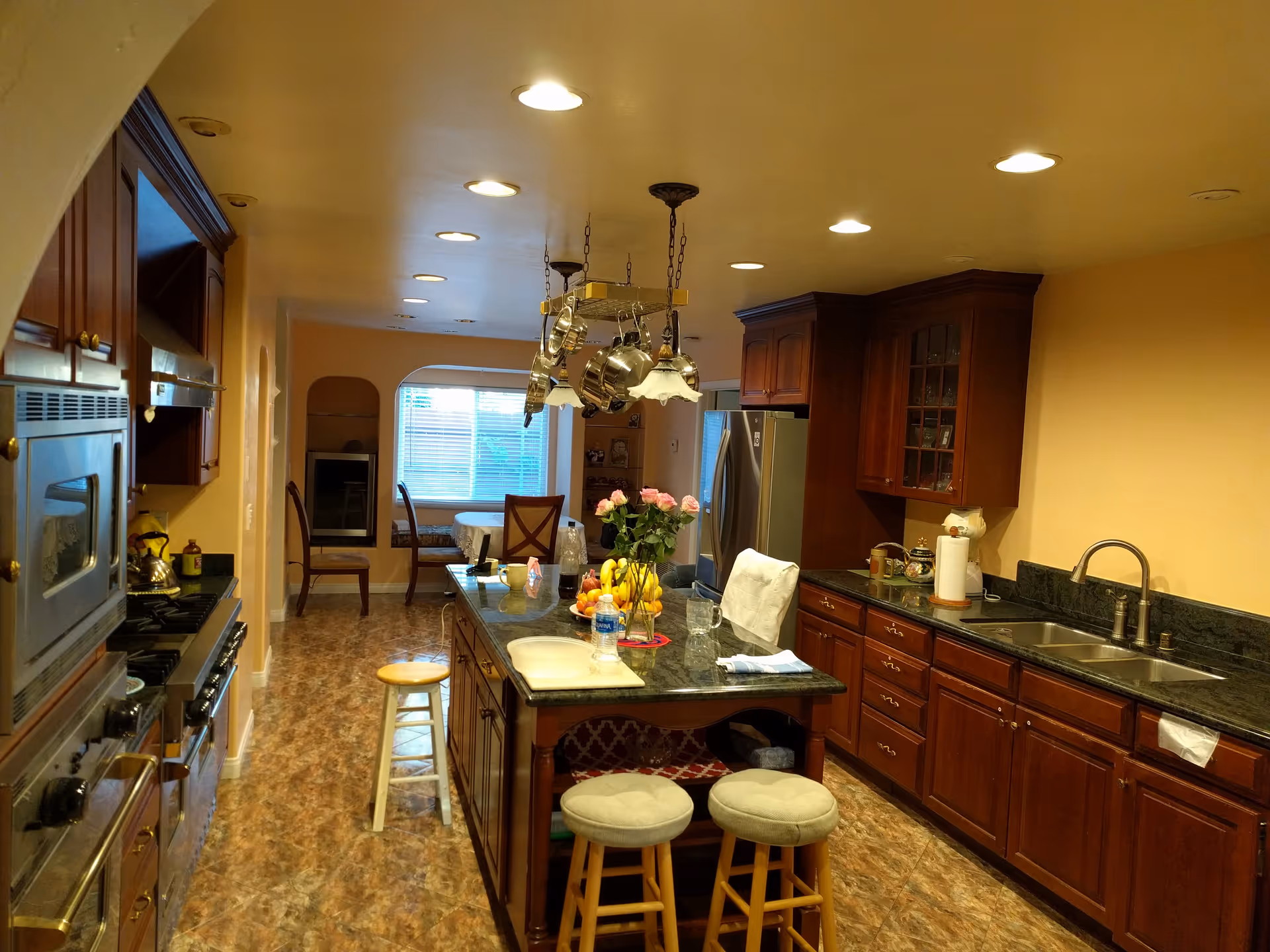 A spacious kitchen with wooden cabinets and a dark granite countertop island in the center. The island has two cushioned stools and a vase with pink roses. Stainless steel appliances, including an oven and refrigerator, are visible. The kitchen opens into a dining area with chairs and a table near a window with blinds.