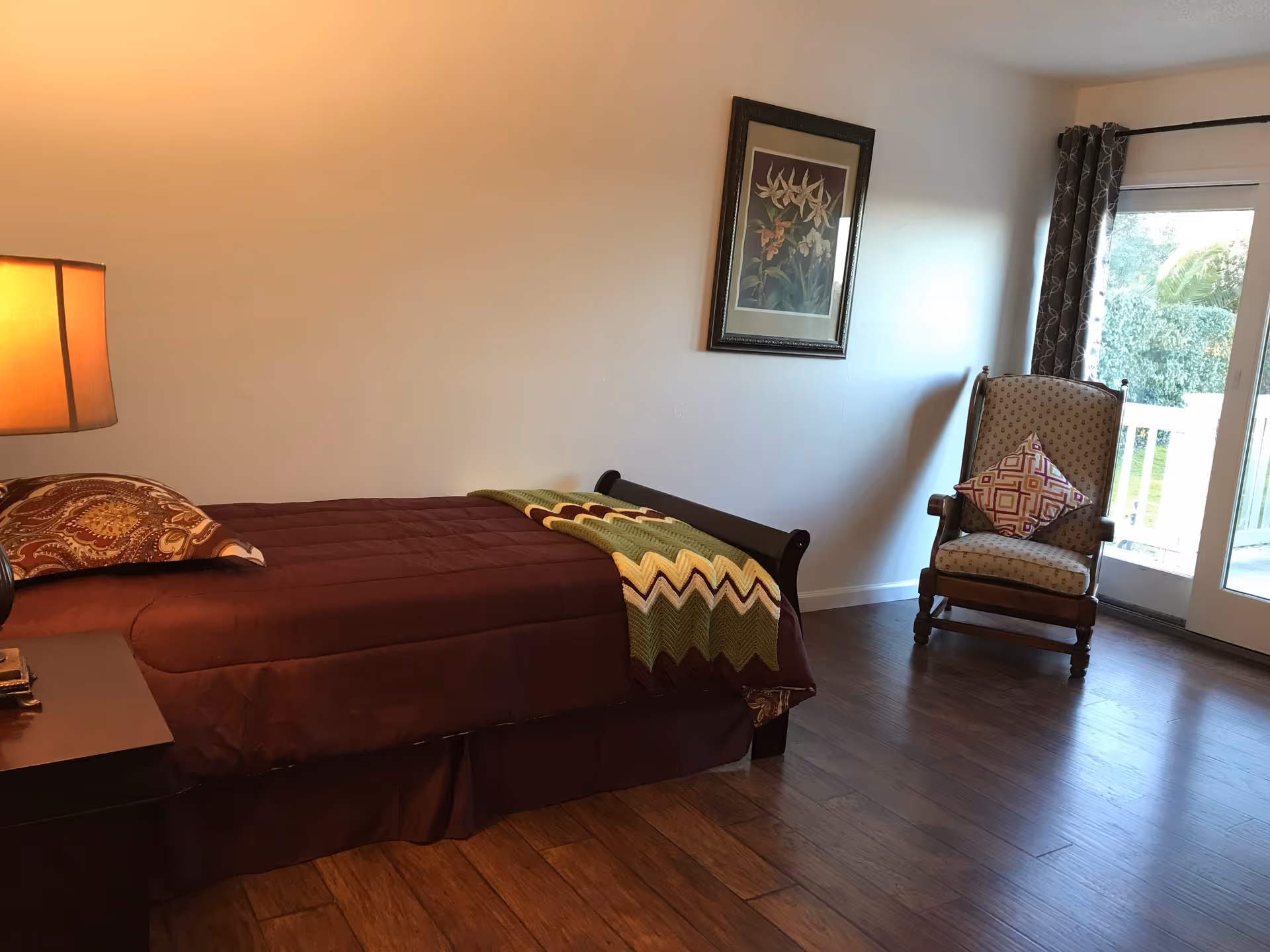 A bedroom with a single bed covered in a brown bedspread and a patterned pillow. A green and yellow zigzag blanket is draped over the foot of the bed. There is a wooden nightstand with a lamp on it next to the bed. A framed floral picture hangs on the wall above the bed. To the right, there is a cushioned armchair with a decorative pillow, positioned near a glass door that opens to an outdoor area with greenery visible outside.