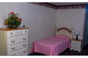 Single-occupancy bedroom with a twin bed covered by a pink bedspread, a white dresser topped with flowers, and a nightstand with a lamp.
