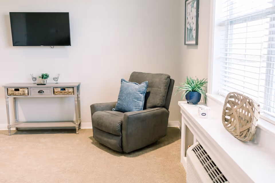 A cozy living room area with a gray recliner chair featuring a blue pillow, a small console table with decorative items underneath a wall-mounted flat screen TV, and a window with white blinds letting in natural light. A white shelf below the window holds a potted plant and a decorative basket.
