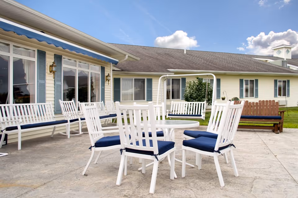 Outdoor patio area at Walden Place with white chairs featuring blue cushions arranged around a white round table. There are additional white benches with blue cushions along the building wall and a white porch swing. The building has cream-colored siding with blue shutters and a blue awning. The sky is partly cloudy.