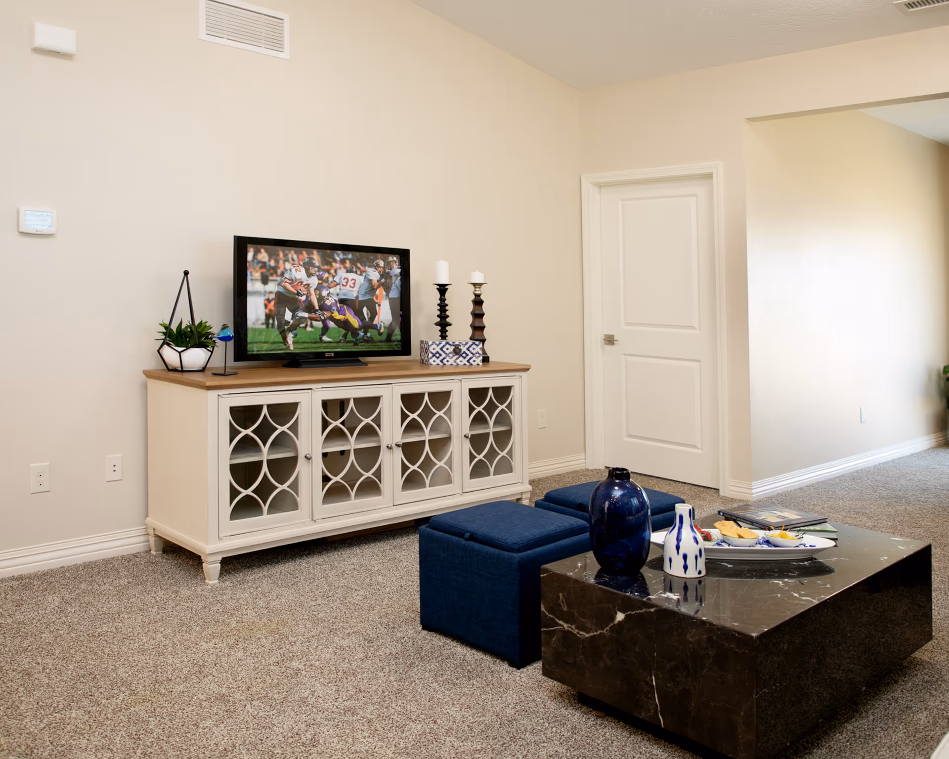 A cozy living room with a TV on a decorative white media console, blue ottomans, and a dark marble coffee table topped with vases and snacks.