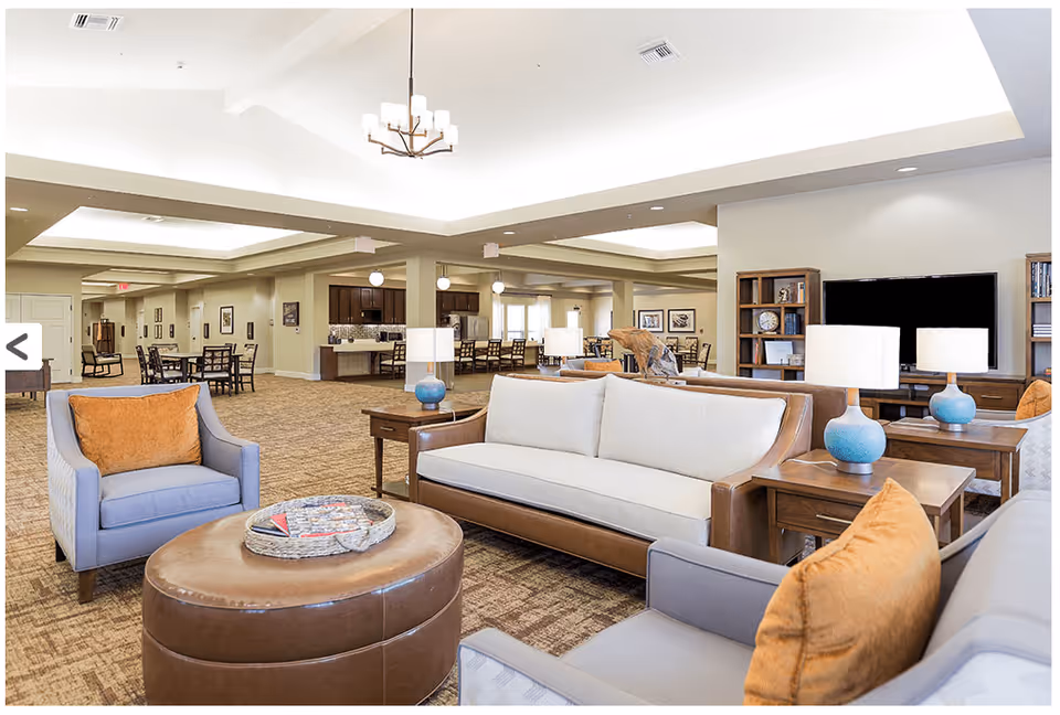 A spacious and well-lit common living area in a senior living facility featuring comfortable seating including sofas and armchairs with orange cushions, a large round leather ottoman with a tray on top, wooden side tables with blue lamps, and a flat-screen TV on a wooden stand. In the background, there are dining tables and chairs, and a kitchen area with pendant lights.