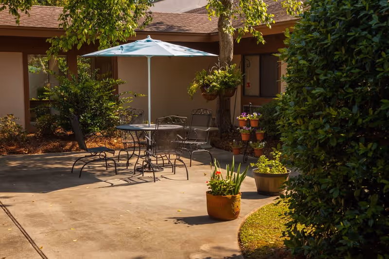 Outdoor patio area with a round metal table and four matching chairs under a light blue umbrella. The patio is surrounded by greenery, including potted plants and bushes, with a building in the background.